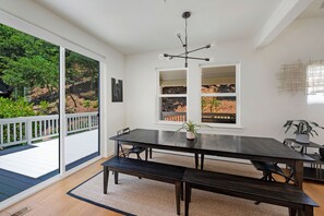 Un espace repas avec une table et des bancs en bois foncé, de grandes fenêtres donnant sur un jardin et un luminaire au plafond.