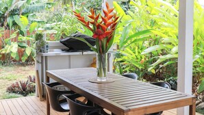 A wooden deck with a table and chairs, a ceiling fan, and a variety of tropical plants in the background. - The Green House (Rarotonga)