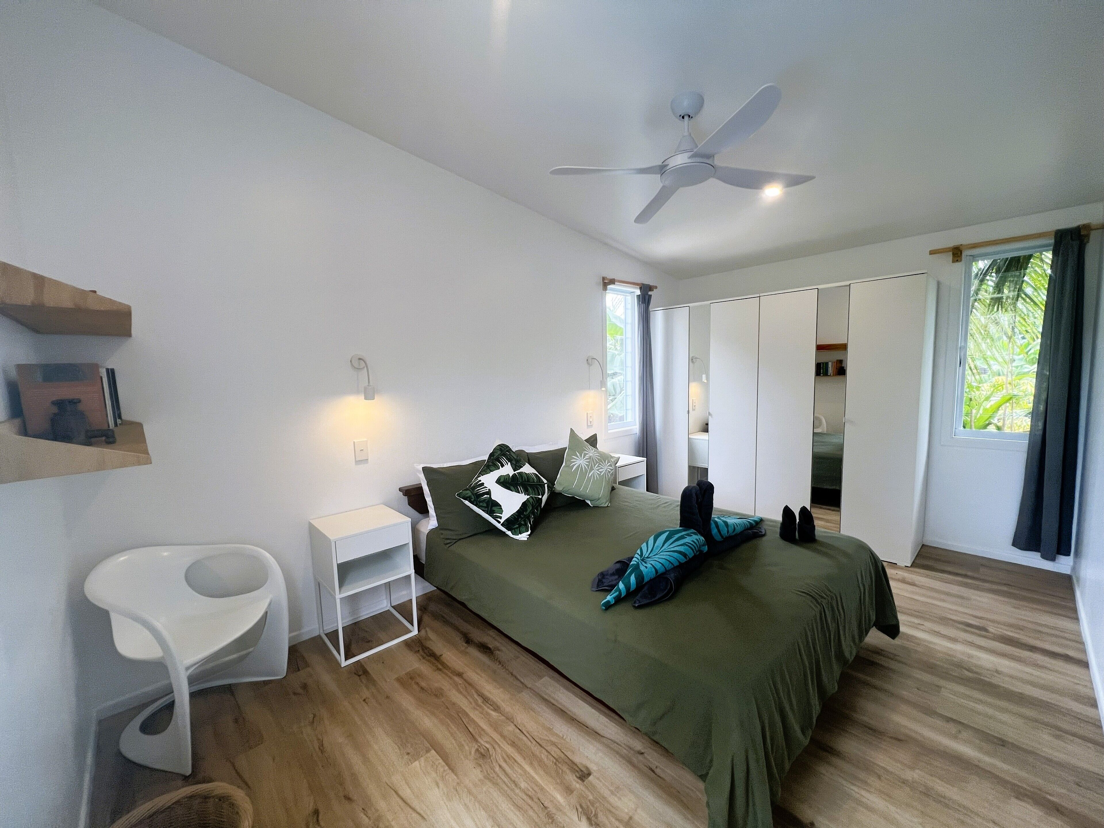A modern bedroom with a bed, a ceiling fan, a nightstand, a white chair, and a window with curtains.