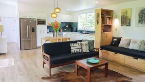A modern kitchen with white cabinets, a stainless steel refrigerator, and a microwave. There is a dining area with a wooden table and chairs, and a living room with a black sofa and a coffee table. - The Green House (Rarotonga)