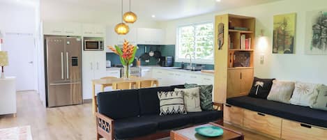 Eine moderne Küche mit weißen Schränken, einem Edelstahlkühlschrank und einer Mikrowelle. Es gibt einen Essbereich mit einem Holztisch und Stühlen sowie ein Wohnzimmer mit einer schwarzen Couch und einem Couchtisch.