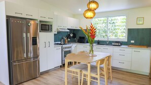 A modern kitchen with white cabinets, stainless steel appliances, a wooden dining table, and a large window. - The Green House (Rarotonga)