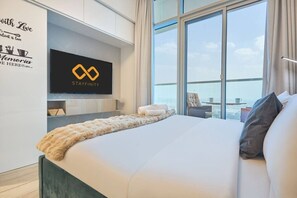 A modern hotel room with a large bed, a TV mounted on the wall, and a view of the city through large windows. - Cozy Marina Studio w/ Balcony (Dubai)