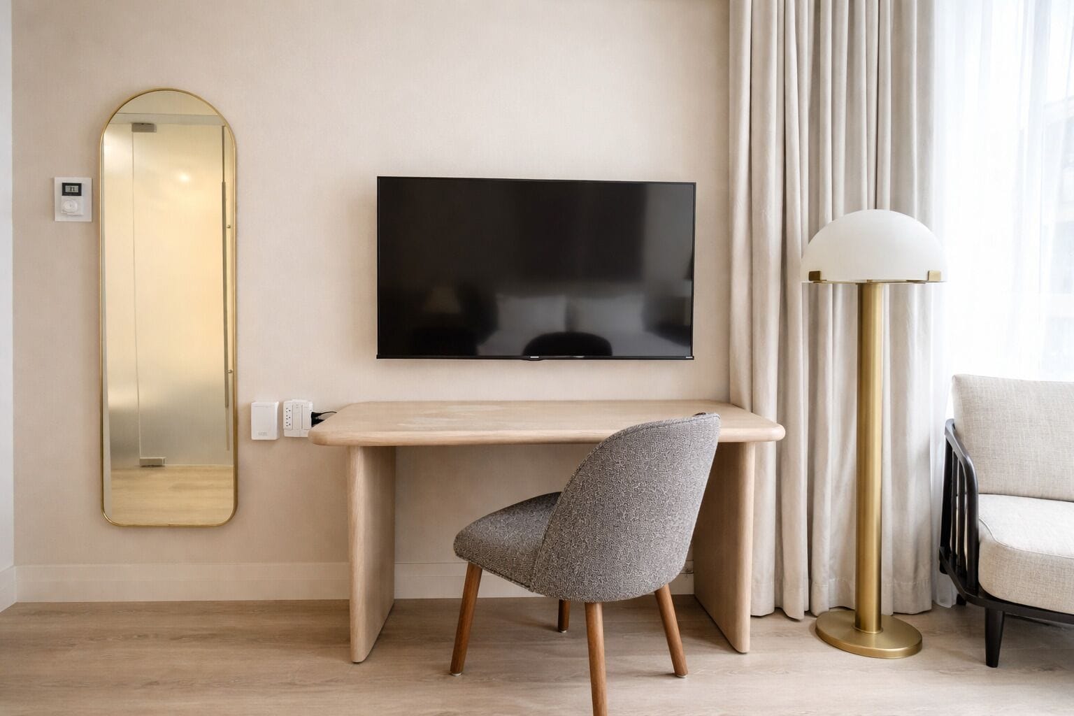 A modern room with a flat-screen TV mounted on the wall, a wooden desk, a grey upholstered chair, a floor lamp, and a sofa.