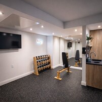 Een moderne, goed verlichte kamer met een wandgemonteerde televisie, een houten wijnrek, fitnessstoelen en een spoelgedeelte.