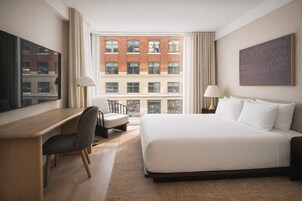 A hotel room with a large bed, a desk, a chair, and a view of a red brick building.