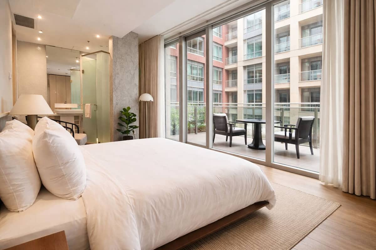 Habitación de hotel con una cama grande, un área de descanso con una mesa y sillas, un amplio ventanal con vistas al exterior del edificio, y un espejo con lámpara.