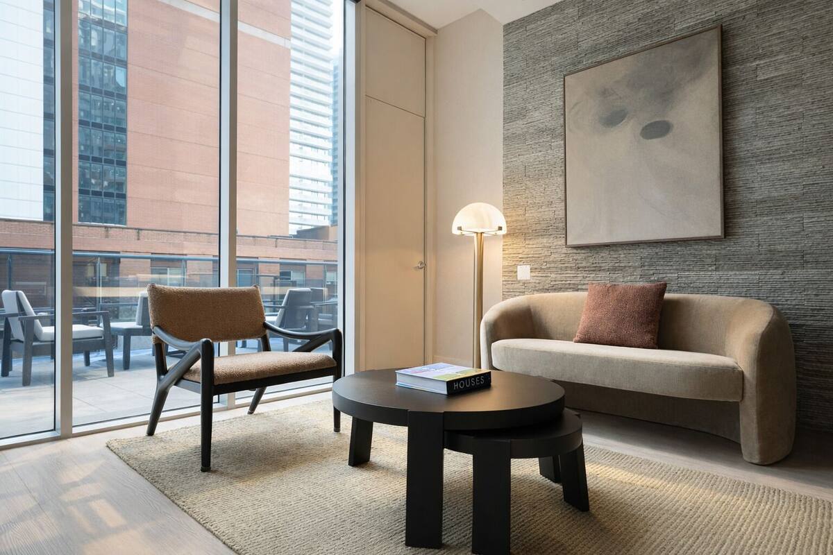 Una habitación de hotel moderna con un ventanal grande, un sofá, una silla, una mesita y una lámpara de pie.