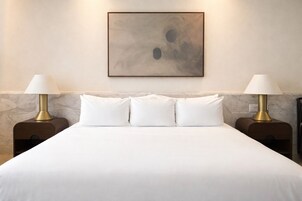Een net opgemaakt bed met witte lakens, met aan weerszijden twee nachtkastjes met lampen, tegen een marmeren muur met daarboven een lijst met een abstract schilderij.