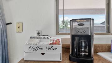 Una cafetera sobre una encimera junto a una caja blanca con el rótulo 'Coffee Bar', con vasos desechables y cápsulas de café.