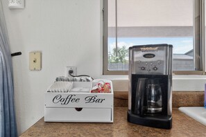 Een koffiezetapparaat op een aanrecht, naast een witte doos met de tekst 'Coffee Bar', met wegwerpbekers en koffiepads.