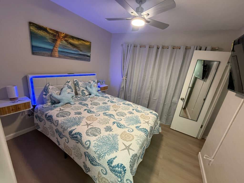 A bedroom with a bed, a ceiling fan, a nightstand, a painting, and a door with a mirror.