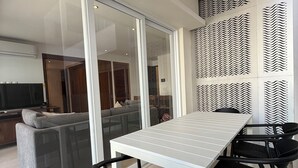 Een moderne eethoek met een witte tafel en zwarte stoelen, een glazen schuifdeur naar een woonkamer met een grijze bank en een televisie.