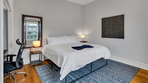 Ein Schlafzimmer mit einem Bett, einem Schreibtisch mit Lampe, einem Sessel, einem gemusterten Teppich und einem Wandbild.