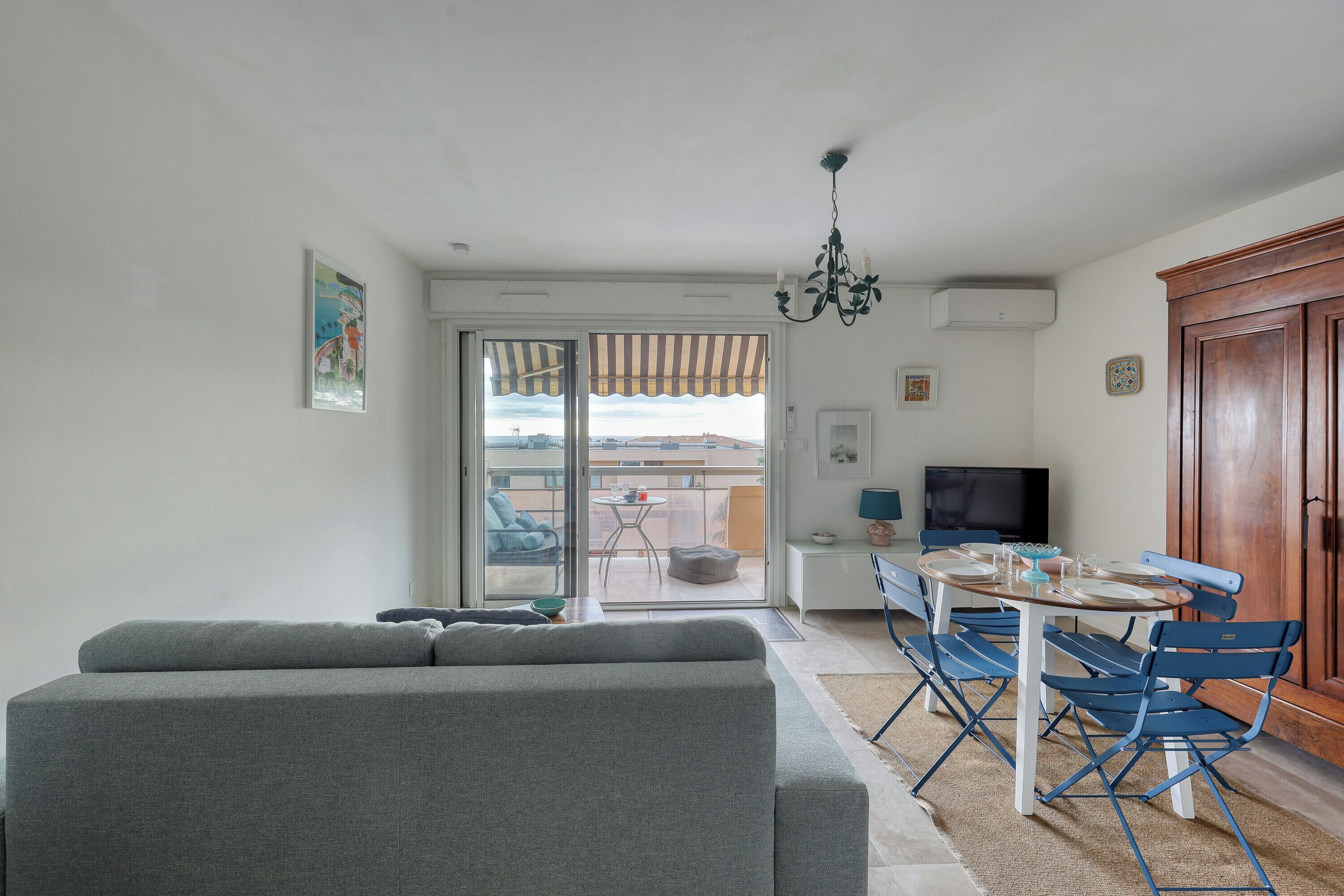 Un soggiorno con un divano grigio, un tavolo da pranzo con sedie blu, una televisione e vista su un balcone esterno attraverso una porta scorrevole in vetro.