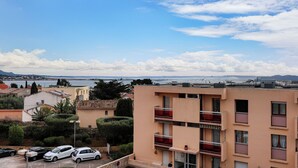 A multi-story building with balconies, a parking lot with cars, and a view of the sea and mountains in the background. - Studio Merle - Welkeys (Bandol)