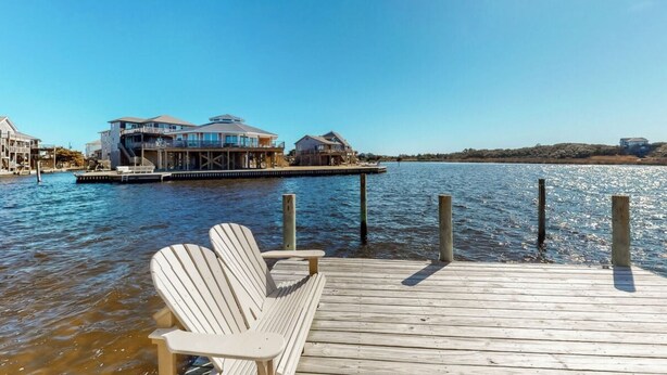 Un muelle de madera con dos sillas Adirondack, una propiedad frente al agua con varios edificios, y un cielo azul despejado.