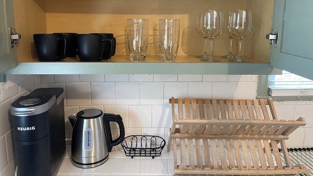 A kitchen with a wooden shelf holding black mugs and glasses, a kettle, and a coffee maker.