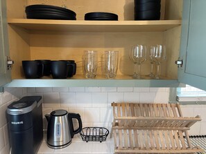 A kitchen with a wooden shelf holding black mugs and glasses, a kettle, and a coffee maker.