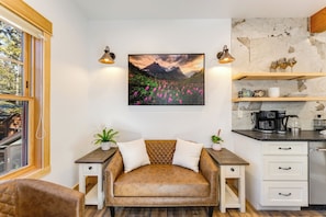 Un salon chaleureux avec un canapé en cuir marron, une table basse en bois et un tableau représentant un paysage de montagne. On y trouve deux tables de chevet avec des lampes, une commode blanche avec des tiroirs et, par la fenêtre, une vue sur la montagne.