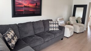 A living room with a grey sofa, a framed sunset picture, and a white armchair.