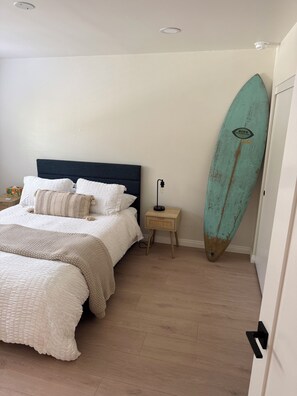 Quarto com cama, mesa de cabeceira, luminária e uma prancha de surf encostada na parede.