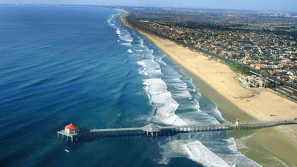 An aerial view of a coastal area with a beach, a pier, and a red-roofed structure.