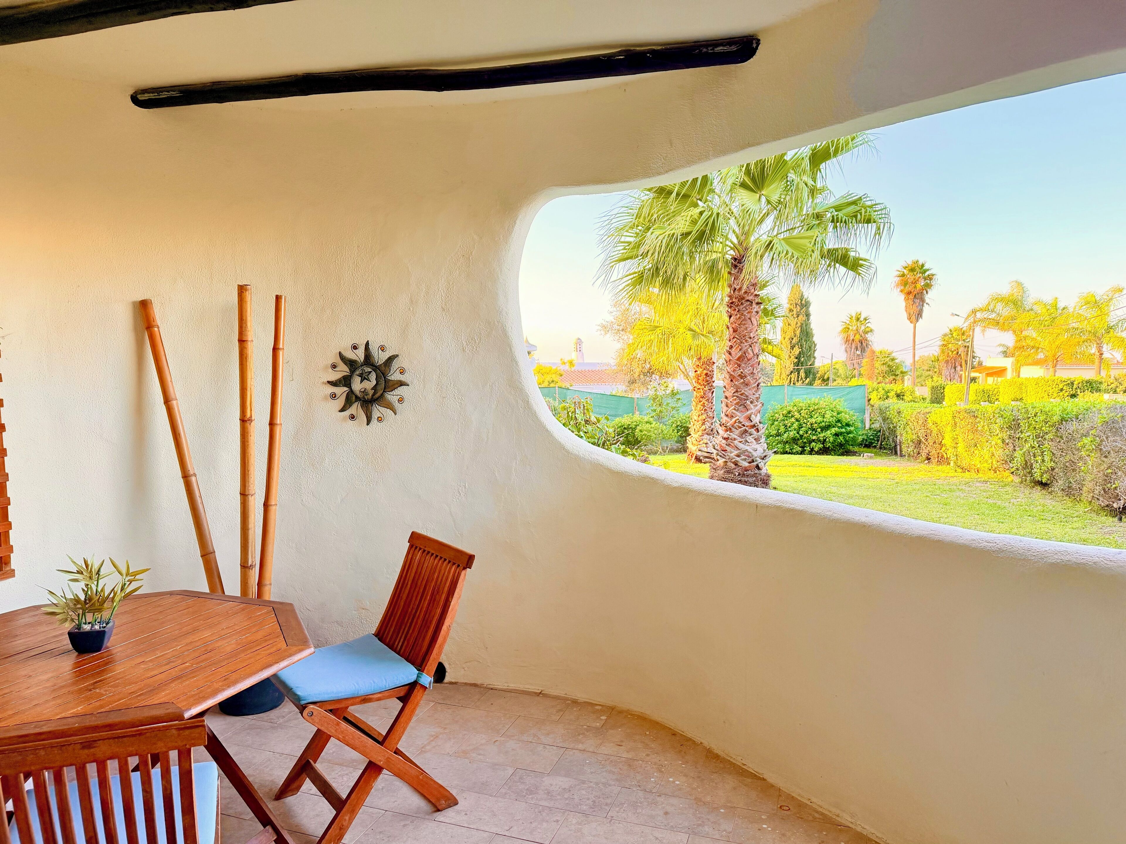 Ein Zimmer mit einem Holztisch, einem Klappstuhl, einem Wanddekor und einem Fenster mit Blick auf einen Garten mit Palmen.