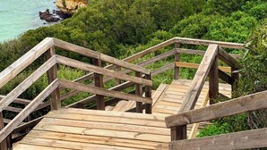 Escadaria de madeira que leva a um mirante com vista para um penhasco costeiro e uma formação rochosa em arco.