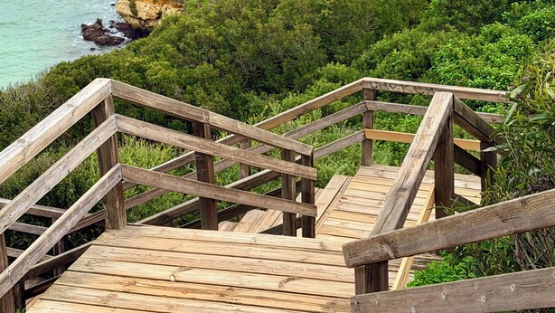 Una escalera de madera conduce a un mirador sobre un acantilado costero con una formación rocosa en forma de arco.