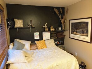 Un dormitorio con cama, almohadas, un estante en la pared con adornos, un cuadro enmarcado y una mesita de noche con lámpara.