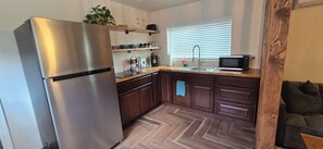 A compact kitchen with a stainless steel refrigerator, wooden cabinets, a microwave, and a window with blinds.