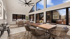 Un espace intérieur spacieux avec une grande table en bois entourée de chaises en osier, de grandes fenêtres donnant sur l’extérieur et un ventilateur de plafond.