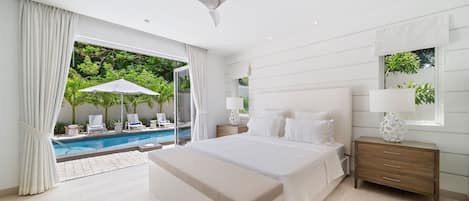 A modern bedroom with a large bed, a nightstand, a ceiling fan, and a view of a pool and greenery outside.