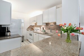 A modern kitchen with white cabinets, a granite countertop, a coffee maker, and a vase with tulips. - Oceanfront and beach front - Menehune Shores 411 (Kihei)