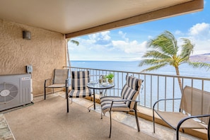 A balcony with a Mitsubishi air conditioning unit, two striped chairs, a small round table with a plant, and a view of the sea and palm tree. - Oceanfront and beach front - Menehune Shores 411 (Kihei)