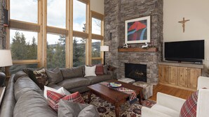 A cozy living room with a stone fireplace, a large window, and a television. - Ski In/Out | Black Eagle Base-Inn | Big Sky Resort (Big Sky)