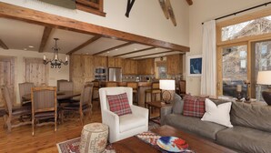 A spacious living room with wooden floors, a dining area with a wooden table and chairs, a kitchen with wooden cabinets, and a large window with curtains. - Ski In/Out | Black Eagle Base-Inn | Big Sky Resort (Big Sky)