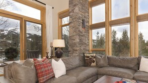 A cozy living room with a stone fireplace, large windows with mountain views, and a sectional sofa with various pillows. - Ski In/Out | Black Eagle Base-Inn | Big Sky Resort (Big Sky)