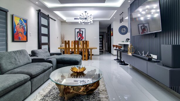 Ein modernes Wohnzimmer mit einem grauen Sofa, einem Glastisch mit Holzfuß, einem Essbereich mit Holztisch und -stühlen sowie einem an der Wand befestigten Fernseher.