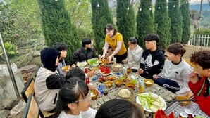 A group of people seated around a table with various dishes, in an outdoor setting with trees and a cloudy sky.