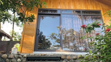 Una cabaña de madera con grandes puertas correderas de vidrio, un muro de piedra en el frente y abundante vegetación alrededor.