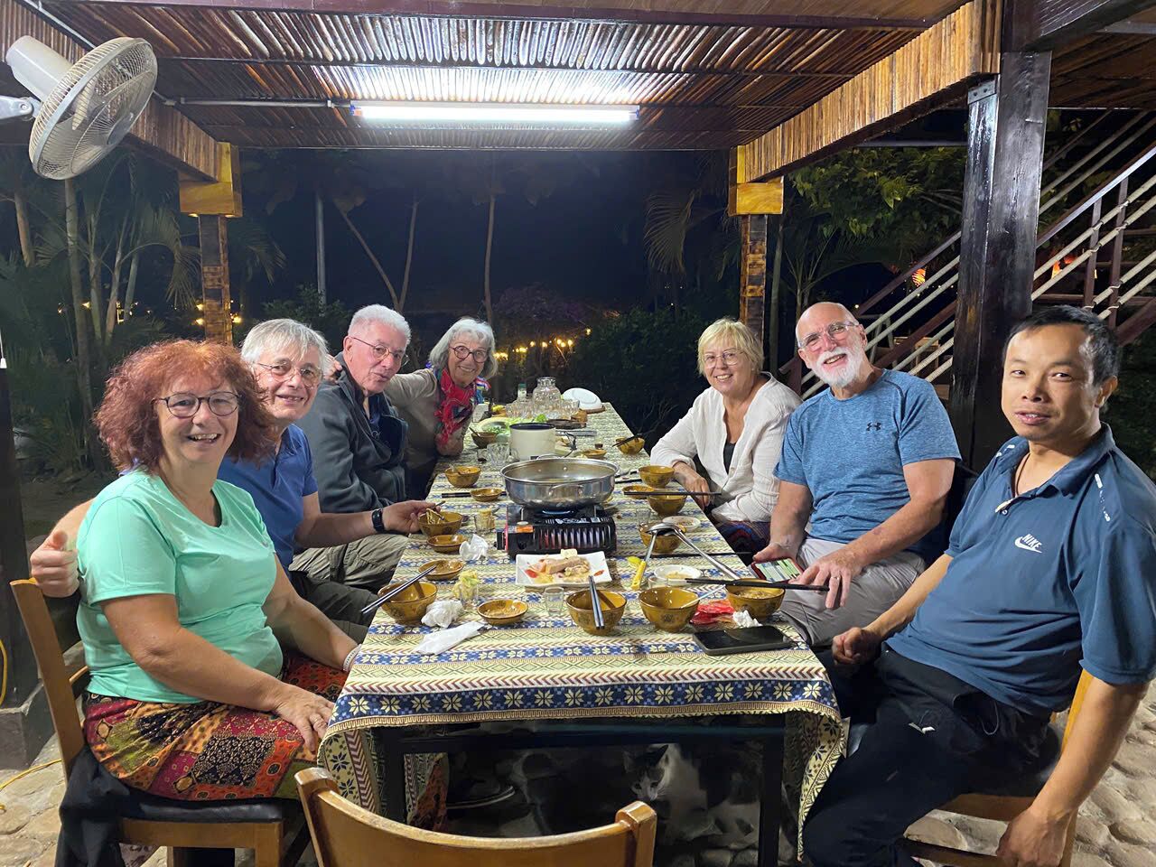 Un grupo de personas sentadas alrededor de una mesa con mantel estampado, un abanico y una parrilla en el centro.