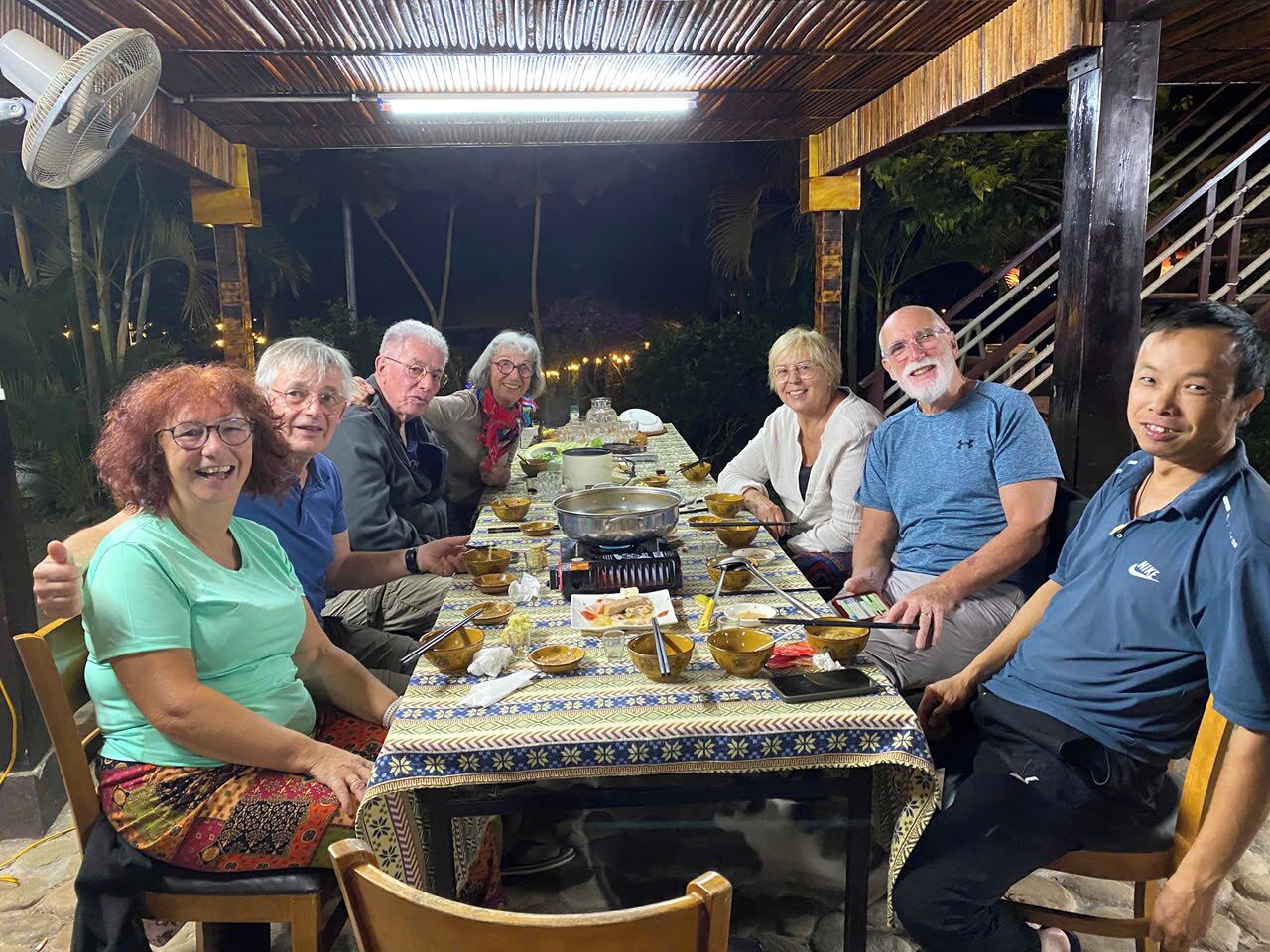 Un groupe de personnes est assis autour d’une table, où sont disposés divers plats, et un réchaud portatif se trouve à proximité. L’ensemble est installé dans un espace extérieur abrité, équipé d’un ventilateur et décoré de guirlandes lumineuses.