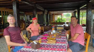 Een overdekte eethoek met een lange tafel gedekt voor een maaltijd, omgeven door houten pilaren en een uitzicht op groen in de achtergrond.