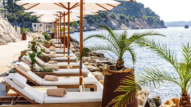 Een rij houten ligbedden met handdoeken, een grote potplant en parasols aan een strandboulevard.