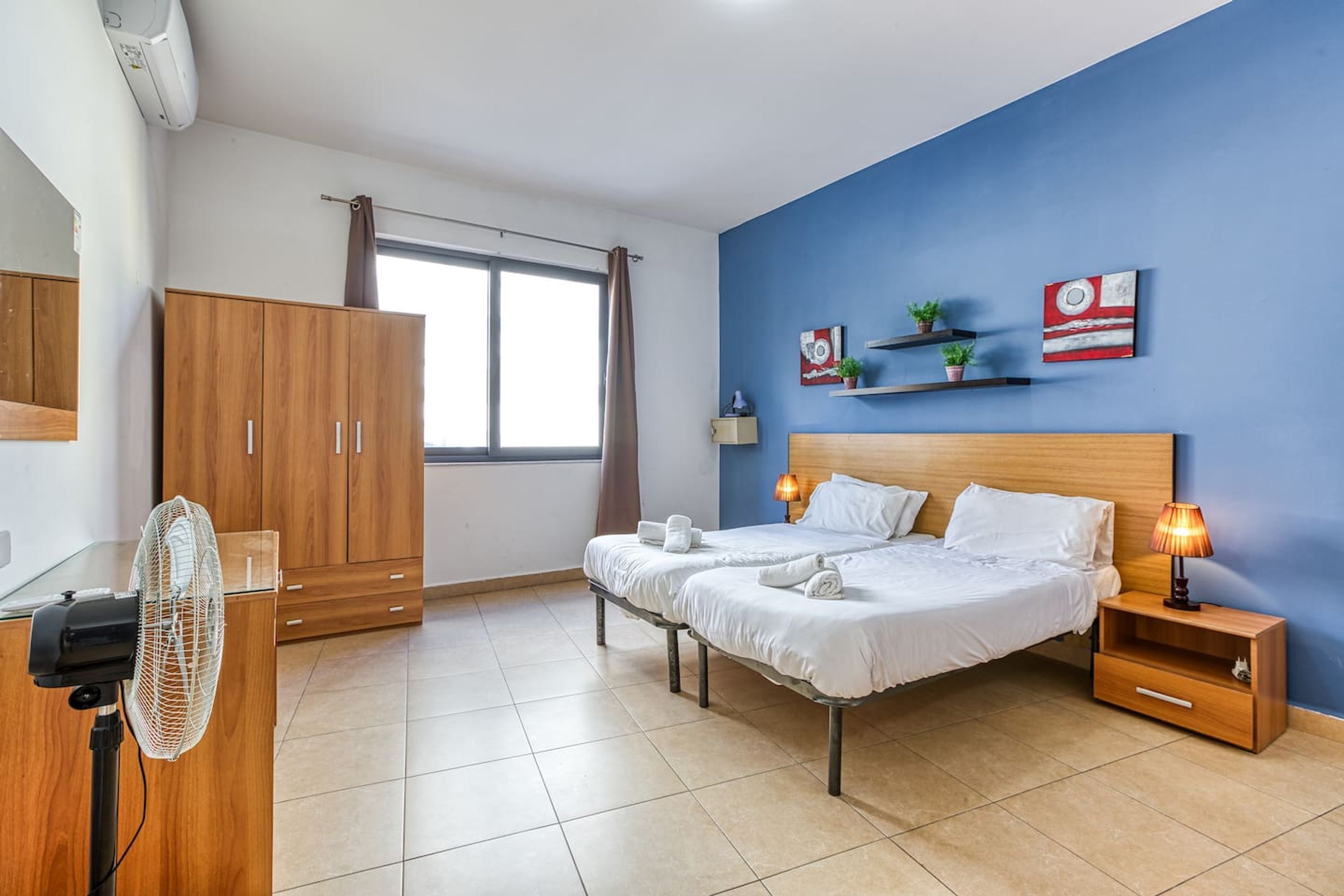 Una habitación de hotel con cama, dos mesitas de noche, un tocador, un ventilador, armario, ventana con cortinas y adornos en la pared.
