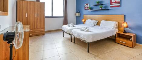 Una habitación de hotel con cama, dos mesitas de noche, un tocador, un ventilador, armario, ventana con cortinas y adornos en la pared.