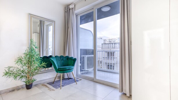 Een moderne woonkamer met een grote schuifglazen deur die leidt naar een balkon, een groene stoel, een potplant en een spiegel aan de muur.