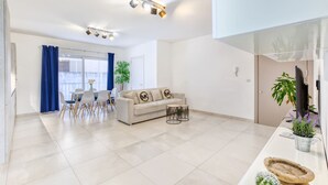 Een moderne woonkamer met een bank, eettafel en stoelen. Er is een keukenhoek met een aanrecht en planten.